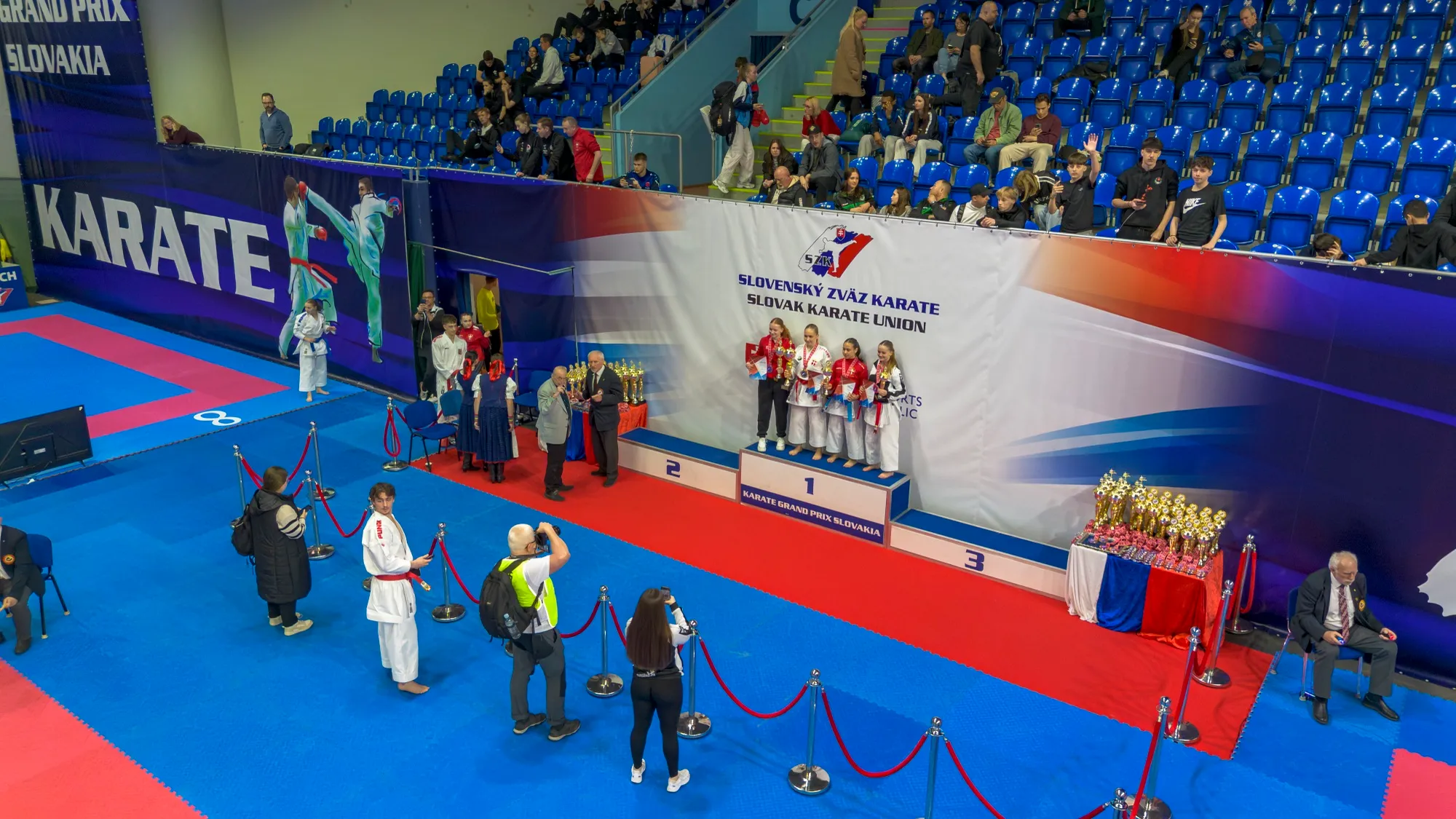 Medal ceremony at 46. Grand Prix Slovakia 2026 in Bratislava, with the placed competitors standing on the numbered podium in front of the Slovak Karate Union backdrop and trophies waiting on the table beside them.